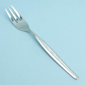 May include: A stainless steel fork with three tines and a long, slender handle. The handle features a subtle, etched design. The fork is set against a light blue background, highlighting its polished surface. Ideal for dining.