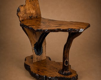 May include: A side table constructed from natural wood slabs with a live edge finish. The table features a vertical back support, a horizontal tabletop, and a single curved branch-like leg, all connected by black metal hardware.
