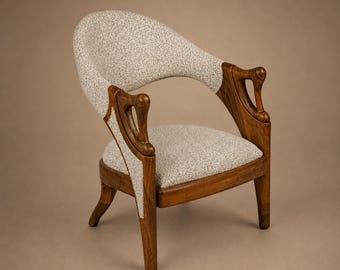 Puede incluir: Un sillón con estructura de madera marrón oscuro, con reposabrazos y patas curvados y esculpidos. El asiento y el respaldo redondeado están tapizados en una tela texturizada con motas de color gris claro y blanco, contrastando con la madera.