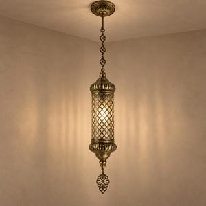 May include: A cylindrical pendant light fixture with an antique brass finish, featuring a central glass panel with a diamond-shaped metal lattice overlay. The lamp hangs from a decorative chain and includes a pointed, ornate metal finial at the base.
