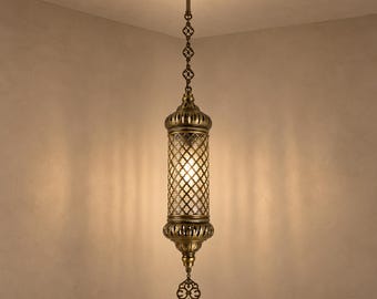 May include: A cylindrical pendant light fixture with an antique brass finish, featuring a central glass panel with a diamond-shaped metal lattice overlay. The lamp hangs from a decorative chain and includes a pointed, ornate metal finial at the base.