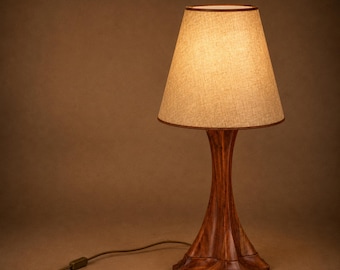 May include: A table lamp featuring a polished wooden base with a curved, fluted design and a tapered, light beige fabric lampshade. The lamp emits a warm, soft glow and includes a brown electrical cord with an inline switch attached.