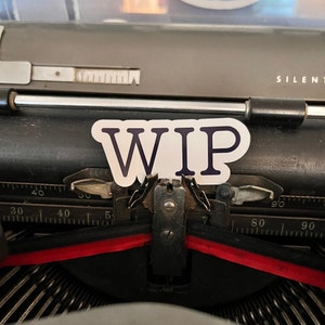 May include: A close-up of a vintage typewriter with a white sticker that says "WIP" on it. The typewriter is black and has a red ribbon.