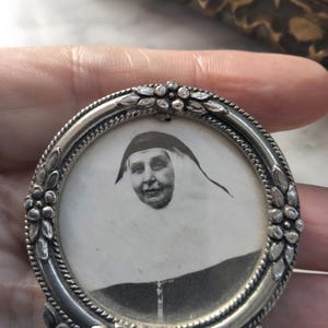 Puede incluir: Un marco de fotos ovalado de plata con un diseño floral. El marco contiene una foto en blanco y negro de una mujer con un cubrecabezas blanco.
