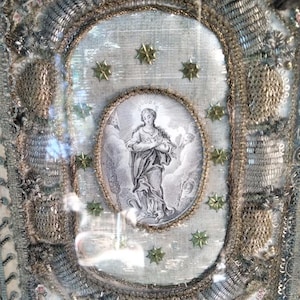 Puede incluir: Una imagen bordada enmarcada de la Virgen María con halo y corona, rodeada de estrellas doradas sobre fondo blanco. El marco es dorado con un borde decorativo.