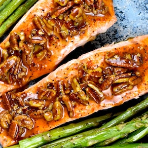Könnte beinhalten: Zwei Lachsfilets, belegt mit Pekannüssen und Glasur, serviert mit grünen Spargel. Der Lachs hat eine rot-orange Farbe, die Pekannüsse sind braun und beige. Der Spargel ist hellgrün.