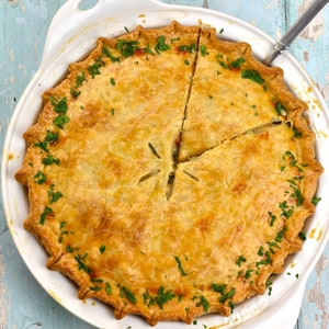 Può includere: Una torta di pollo dorata con una crosta sfogliata, guarnita con prezzemolo fresco, è tagliata in tre fette e servita in una tortiera bianca.