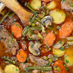 Könnte beinhalten: Eine Nahaufnahme eines Topfes mit herzhaftem Eintopf mit Rindfleisch, Kartoffeln, Karotten, grünen Bohnen, Pilzen und Erbsen in einer reichhaltigen braunen Brühe. Der Eintopf ist mit frischer Petersilie garniert.