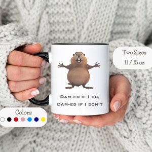 Könnte beinhalten: Eine weiße Keramiktasse mit schwarzem Rand und Henkel. Die Tasse zeigt ein Cartoon-Biber mit dem Text "DAM-ED IF I DO, DAM-ED IF I DON'T". Die Tasse ist in zwei Größen erhältlich: 11 oz und 15 oz.