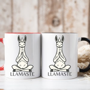 May include: Two white ceramic mugs with black and red accents. One mug has a black handle and the other has a red handle. Both mugs feature a black and white illustration of a llama in a yoga pose with the text "LLAMASTE" printed below.