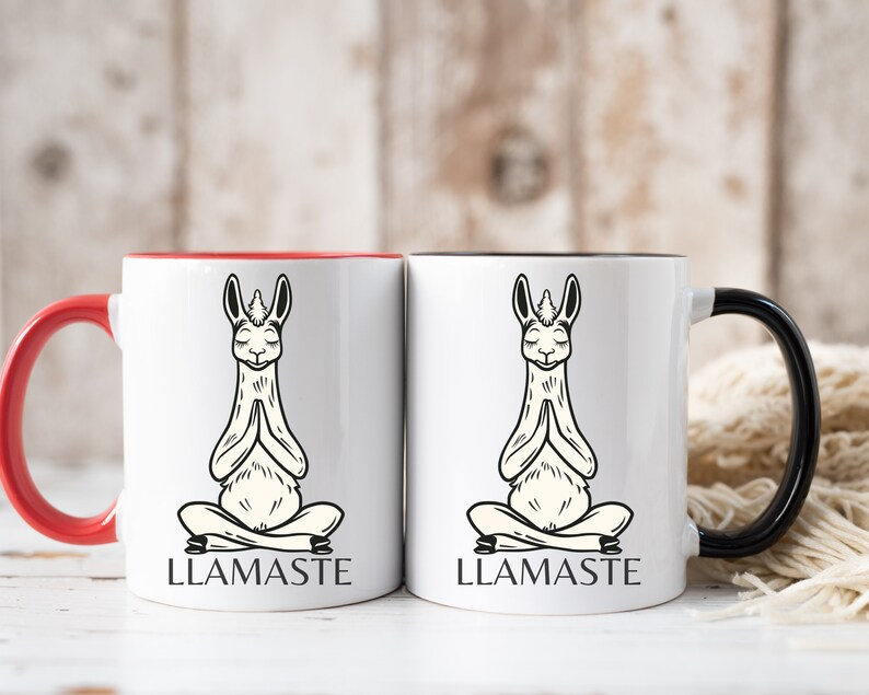 May include: Two white ceramic mugs with black and red accents. One mug has a black handle and the other has a red handle. Both mugs feature a black and white illustration of a llama in a yoga pose with the text "LLAMASTE" printed below.