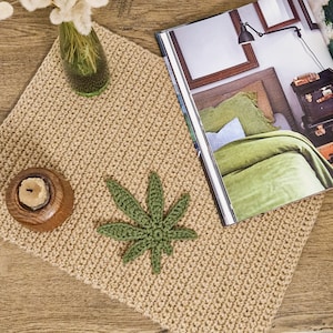 Könnte beinhalten: Ein gehäkelter beigefarbener Teppich mit einem gehäkelten Marihuana-Blattdesign in Grün. Der Teppich liegt auf einer Holzoberfläche mit einer Kerze und einer Glasvase mit getrockneten Blumen.