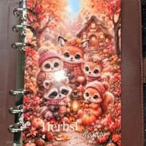 Könnte beinhalten: Ein brauner Ringbuchplaner mit einer herbstlichen Waldszene auf dem Einband. Die Illustration zeigt einen Waschbären, eine Eule, einen Fuchs und ein Eichhörnchen mit Wintermützen und Schals, umgeben von Kürbissen, Laub und einer kleinen Hütte.