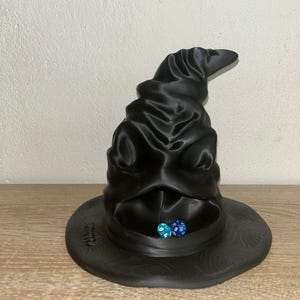 Könnte beinhalten: Ein schwarzer, 3D-gedruckter Sprechender Hut mit breiter Krempe und strukturierten Details. Zwei Würfel, einer blau und einer türkis, liegen im Hut. Der Hut ist eine Requisite aus der Harry-Potter-Reihe.