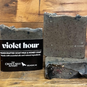 May include: Three bars of handcrafted goat milk and honey soap. The soap is a dark gray color with a textured surface. One bar has a black label that reads "violet hour" and "The Crow's Nest Apiary, Belmont, MI."