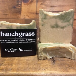 Puede incluir: Barras de jabón artesanales de leche de cabra y miel con aroma a hierba de playa. Las barras son una mezcla de beige y verde, con el texto "beachgrass" en la etiqueta negra. La etiqueta también indica que el jabón está hecho con aceites esenciales e ingredientes naturales.