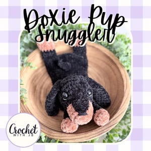 May include: A black and tan crocheted dachshund toy with large dark eyes, positioned inside a wooden bowl. The text Doxie Pup Snuggler is displayed at the top, and a circular logo with the text Crochet With JB is in the bottom corner.