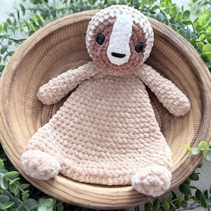 May include: A handmade crochet security blanket featuring a sloth design. The sloth has a brown and white head with black eyes and a white stripe. The blanket is a soft, light brown color, with arms and feet. The blanket is in a wooden bowl.