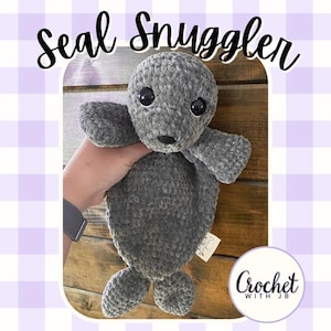 Könnte beinhalten: Ein graues Häkel-Robben-Stofftier mit weicher, strukturierter Garnoberfläche, schwarzen Kunststoff-Sicherheitsaugen und einer kleinen, schwarz gestickten Nase. Der Text "seal Snuggler" steht oben und das Logo "Crochet WITH JB" unten.