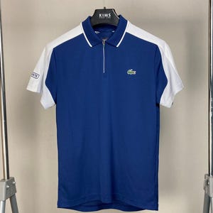 Może przedstawiać: Koszulka polo z krótkim rękawem w kolorze niebieskim i białym, z zamkiem błyskawicznym i zielonym logo krokodyla na piersi. Koszulka ma białe paski po bokach.