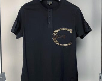 May include: Black short-sleeved shirt with a Henley neckline and a pocket. The pocket features the word "cavalli" in gold-colored embellishments. The shirt has buttoned cuffs and is hanging on a clothing rack.
