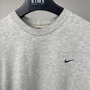 Puede incluir: Una sudadera gris jaspeado con cuello redondo y un peque&ntilde;o logotipo negro de Nike en la parte superior derecha del pecho. La sudadera est&aacute; colgada de una percha negra.