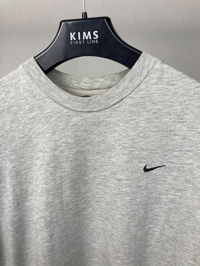 Puede incluir: Una sudadera gris jaspeado con cuello redondo y un peque&ntilde;o logotipo negro de Nike en la parte superior derecha del pecho. La sudadera est&aacute; colgada de una percha negra.