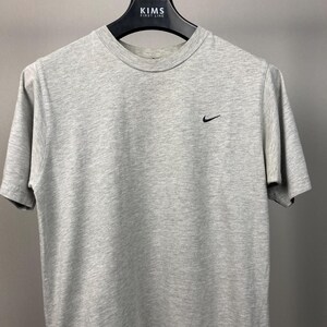 Puede incluir: Una camiseta gris jaspeada de manga corta con un peque&ntilde;o logotipo negro de Nike en el pecho izquierdo. La camiseta est&aacute; colgada de una percha negra con el texto "KIMS FIRST LINE".