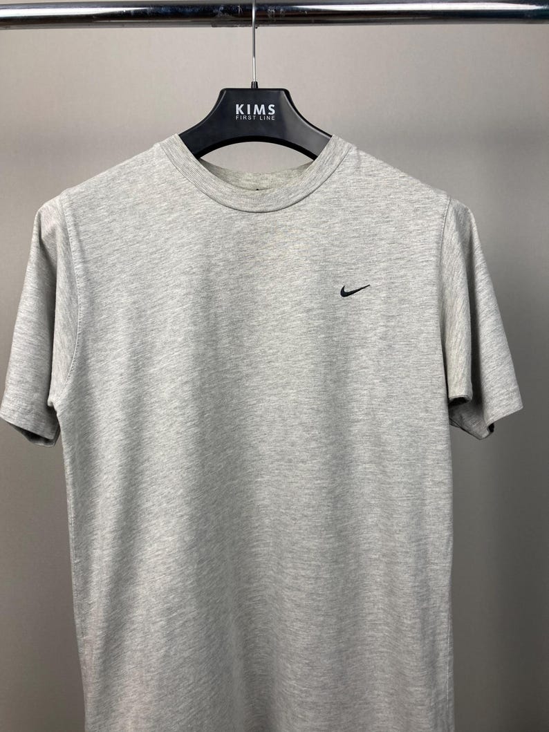 Puede incluir: Una camiseta gris jaspeada de manga corta con un peque&ntilde;o logotipo negro de Nike en el pecho izquierdo. La camiseta est&aacute; colgada de una percha negra con el texto "KIMS FIRST LINE".