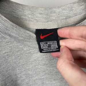 Puede incluir: Primer plano de un cuello de camiseta gris con una etiqueta negra con el logotipo rojo de Nike. La etiqueta incluye medidas de talla y altura, junto con el texto "Made in Greece" y "Fabriqu&eacute; en Gr&egrave;ce".