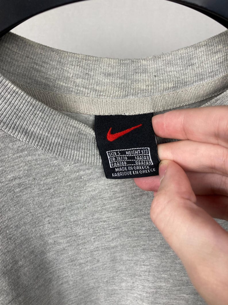Puede incluir: Primer plano de un cuello de camiseta gris con una etiqueta negra con el logotipo rojo de Nike. La etiqueta incluye medidas de talla y altura, junto con el texto "Made in Greece" y "Fabriqu&eacute; en Gr&egrave;ce".