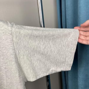 Puede incluir: Primer plano de una manga de camiseta gris claro. La manga est&aacute; hecha de una tela suave y texturizada y tiene un ajuste amplio y relajado. La manga est&aacute; sujeta por una mano, mostrando la forma y la ca&iacute;da de la prenda.
