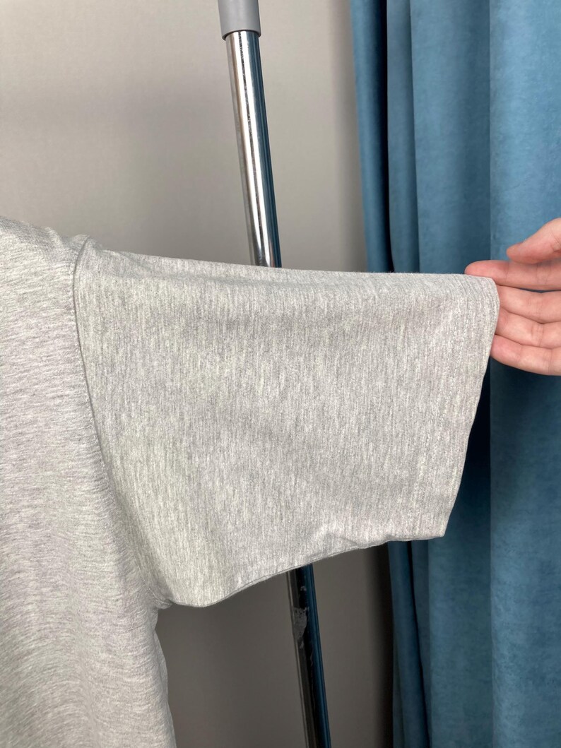 Puede incluir: Primer plano de una manga de camiseta gris claro. La manga est&aacute; hecha de una tela suave y texturizada y tiene un ajuste amplio y relajado. La manga est&aacute; sujeta por una mano, mostrando la forma y la ca&iacute;da de la prenda.