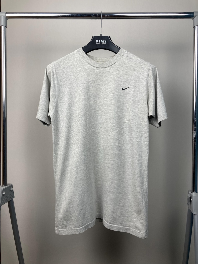 Puede incluir: Una camiseta gris jaspeada de manga corta con un peque&ntilde;o logotipo negro de Nike en el pecho izquierdo. La camiseta est&aacute; colgada de una percha negra con el texto "KIMS" en la parte superior. La camiseta est&aacute; hecha de un material suave y c&oacute;modo.