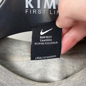 Puede incluir: Primer plano de una camiseta gris con una etiqueta negra. La etiqueta presenta un logotipo blanco de Nike y texto, incluyendo "RN#56323" y "CA#05553". La camiseta est&aacute; colgada en una percha negra. El texto "KIM FIRST LIN" es visible en la percha.