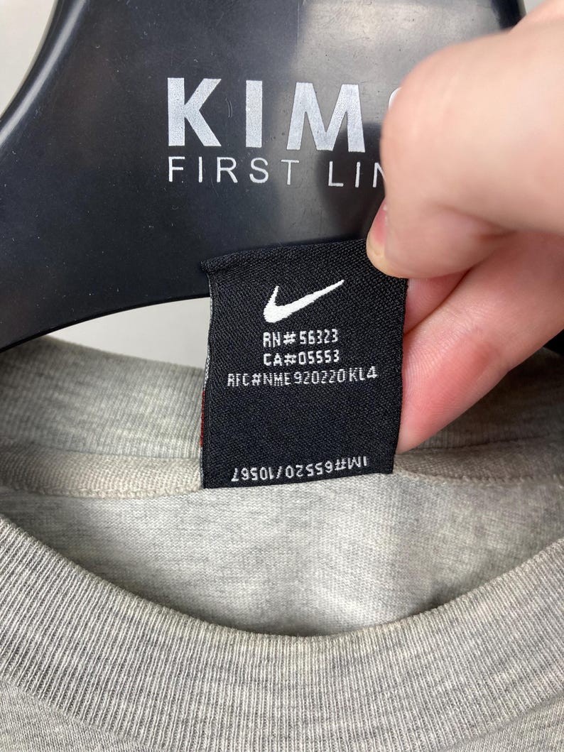 Puede incluir: Primer plano de una camiseta gris con una etiqueta negra. La etiqueta presenta un logotipo blanco de Nike y texto, incluyendo "RN#56323" y "CA#05553". La camiseta est&aacute; colgada en una percha negra. El texto "KIM FIRST LIN" es visible en la percha.