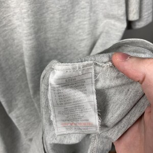 Puede incluir: Primer plano de una camiseta gris con una etiqueta. La etiqueta tiene texto en varios idiomas, incluido el espa&ntilde;ol, y detalla la composici&oacute;n de la tela. La camiseta est&aacute; hecha en Grecia.