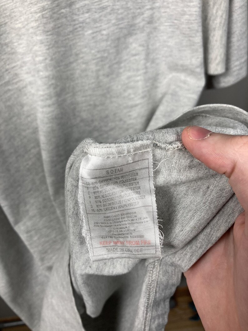 Puede incluir: Primer plano de una camiseta gris con una etiqueta. La etiqueta tiene texto en varios idiomas, incluido el espa&ntilde;ol, y detalla la composici&oacute;n de la tela. La camiseta est&aacute; hecha en Grecia.