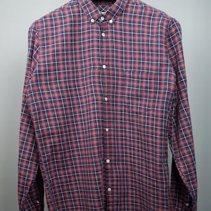 Puede incluir: Una camisa de manga larga con botones y un estampado de cuadros rojos y azules. La camisa tiene un bolsillo en el lado izquierdo y un cuello abotonado.
