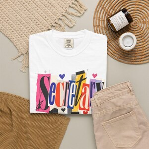 May include: A white short-sleeve t-shirt featuring the word Secretary in a colorful, collage-style font with various paper textures and small heart icons. The shirt has a Comfort Colors brand label at the inner neckline and is displayed flat.