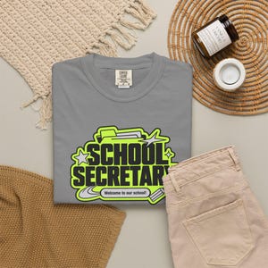 Op de afbeelding: Een grijs t-shirt met een afbeelding met de tekst "SCHOOL SECRETARY" in dikke zwarte letters op een fel neon-groene achtergrond. Het ontwerp bevat een icoon van een map, sterren en een klein lint met "Welcome to our school!"