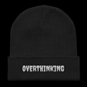 May include: A black ribbed knit beanie featuring a folded cuff. Centered on the front of the cuff is the word OVERTHINKING embroidered in white capital letters with a textured, slightly raised appearance against the dark fabric of the hat.
