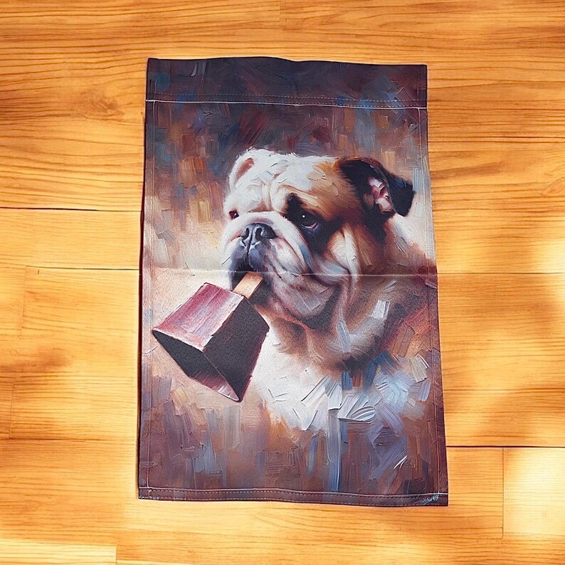 Garden flag featuring a painted portrait of a bulldog holding a maroon cowbell in its mouth, with a warm, abstract background that creates a charming and whimsical atmosphere.