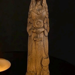 Pode incluir: Uma est&aacute;tua de madeira de uma figura feminina com cabelo comprido, vestindo um vestido longo e segurando uma espada. A est&aacute;tua est&aacute; em p&eacute; sobre uma superf&iacute;cie escura com uma vela acesa &agrave; sua frente.