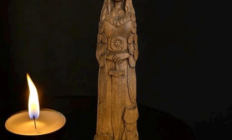 Pode incluir: Uma est&aacute;tua de madeira de uma figura feminina com cabelo comprido, vestindo um vestido longo e segurando uma espada. A est&aacute;tua est&aacute; em p&eacute; sobre uma superf&iacute;cie escura com uma vela acesa &agrave; sua frente.
