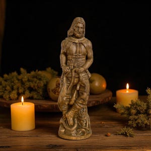 May include: A carved wooden statue of a man with long hair and a beard, dressed in a detailed outfit, holding a weapon and a creature. The statue is in a standing pose, with intricate carvings. The background includes candles and greenery.