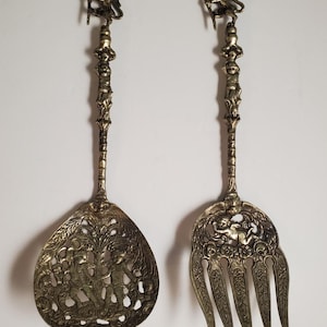 May include: A set of two ornate, gold-toned serving utensils. The serving spoon has a decorative, openwork design with a lion figure at the top of the handle. The serving fork has a similar design with a lion figure at the top of the handle.