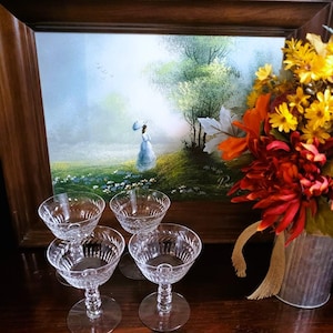 May include: Four clear crystal champagne coupe glasses with etched designs. The glasses are on a dark wooden surface, with a framed painting in the background. A bouquet of colorful flowers in a metal container is also visible.