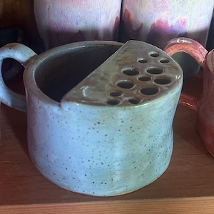 May include: Three ceramic flower frogs with handles, each featuring a semi-circular top with multiple circular openings for stems. The left and right vessels are glazed in shades of orange and pink, while the centre vessel is a light blue-grey.