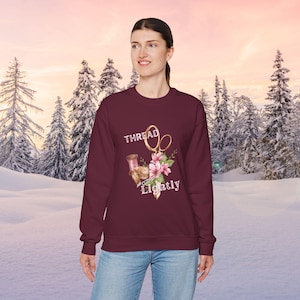 May include: A maroon sweatshirt with the words "THREAD Lightly" and a graphic of gold scissors, pink flowers, and sewing notions. The background features a snowy landscape with evergreen trees.